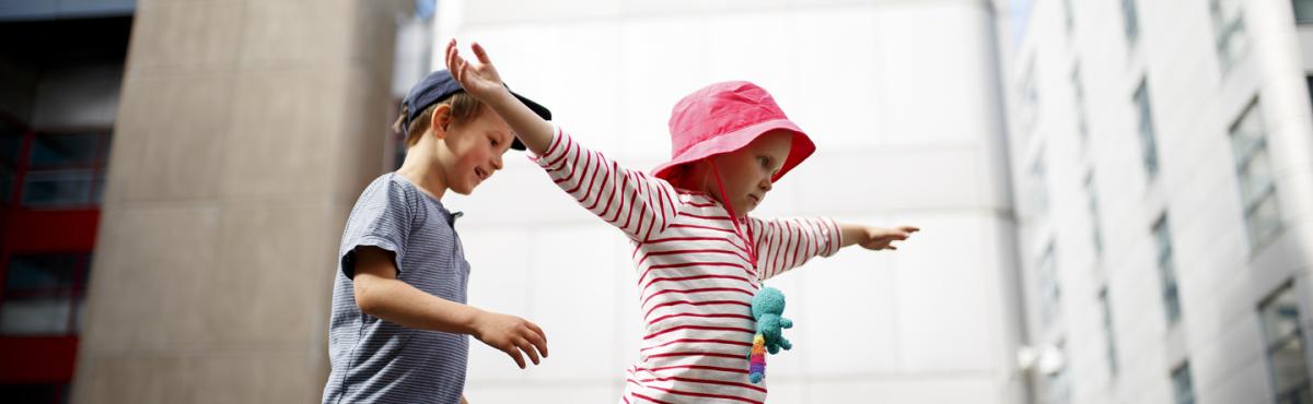 Noin 7-vuotias poika ja tyttö tasapainoilevat pihalla massiivisten kerrostalojen keskellä. Tyttö on levittänyt kätensä, poika tulee perässä. 