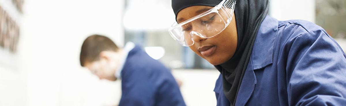 Suojalaseihin ja -takkiin sonnustautunut huivipäinen, teini-ikäinen tyttö katselee keskittyneesti työhönsä luokassa. 