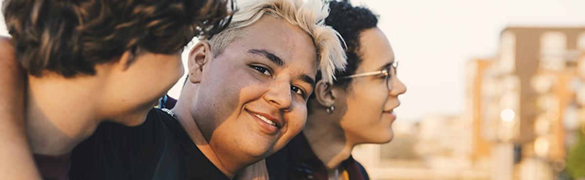 Kolme teini-ikäistä poikaa kävelee kaulakkain kesäillassa kaupunkimiljöössä. Keskimmäinen poika katsoo kameraan hymyillen.