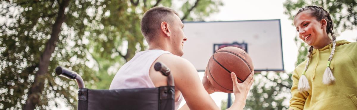 Pyörätuolissa istuva teini-ikäinen poika tarjoaa koripalloa teini-ikäiselle tytölle, joka seisoo hänen vieressään. Taustalla näkyy kori. 