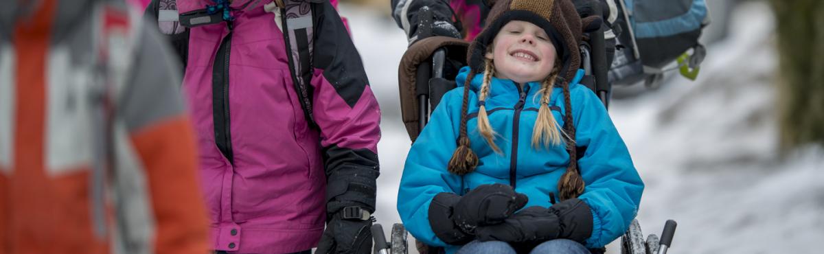 Alakouluikäistä tyttöä työnnetään pyörätuolissa lumisessa maisemassa.