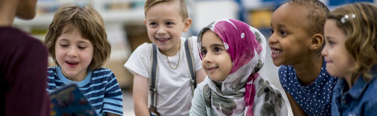 Viiden esikouluikäisen lapsen ryhmä istuu katselemassa kirjan aukeamaa, jota opettaja näyttää. 