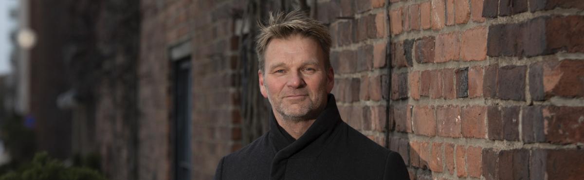 Mies katsoo kameraan, taustalla näkyy tiiliseinää. 