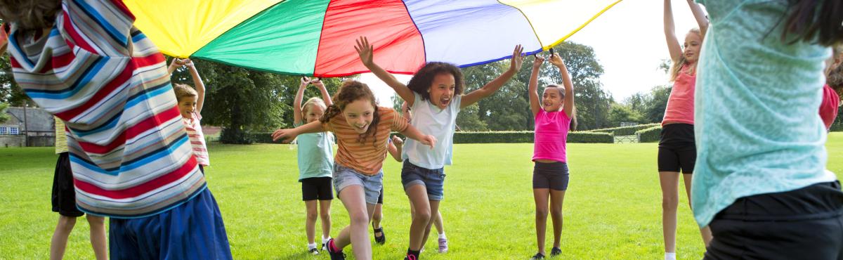 Kaksi tyttöä juoksee värikkään kankaan ali. Muut lapset pitelevät kangasta ylhäällä.