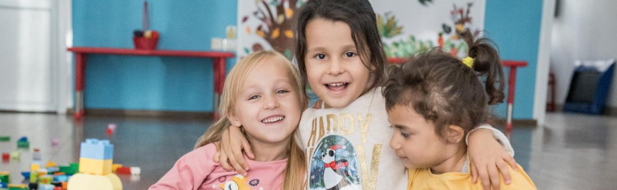 Kolme pientä lasta halaa toisiaan. Lapset istuvat lattialla. Vieressä on Duplo-palikoita.