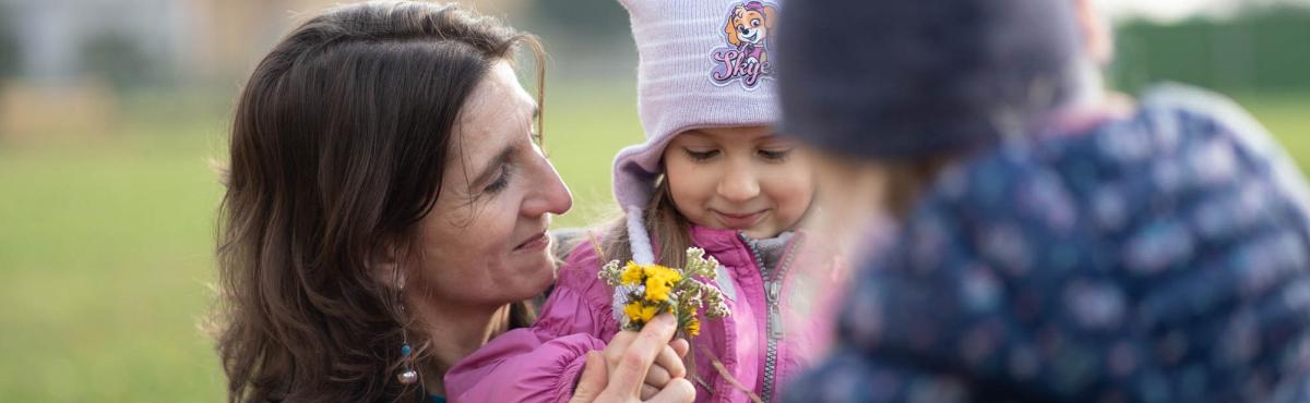 Nainen ja alakouluikäinen, pipoon ja pinkkiin toppatakkiin pukeutunut lapsi pitävät yhdessä keltaisia kukkia kädessään. Taustalla näkyy vihreää niittyä. Etualalla näkyy sumeana toinen lapsi.  