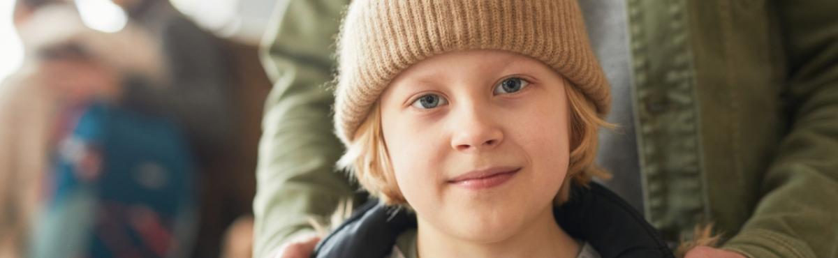 Beigeen pipoon sonnustautunut kouluikäinen poika katsoo kameraan. Pojan taustalla on aikuinen, joka pitää käsiään pojan olkapäillä.