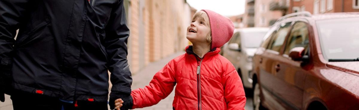 Alakouluikäinen, punaiseen takkiin ja pipoon pukeutunut lapsi pitää aikuista kädestä ja katsoo ylös aikuisen kasvoihin. Taustalla näkyy autoja ja katumaisemaa.