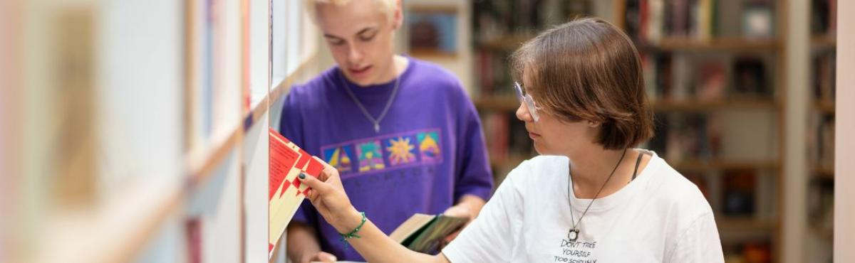 Valkoiseen t-paitaan pkeutunut nuori ottaa hyllystä kirjaa. Taustalla näkyy toinen nuori, joka nojaa kirjahyllyyn.