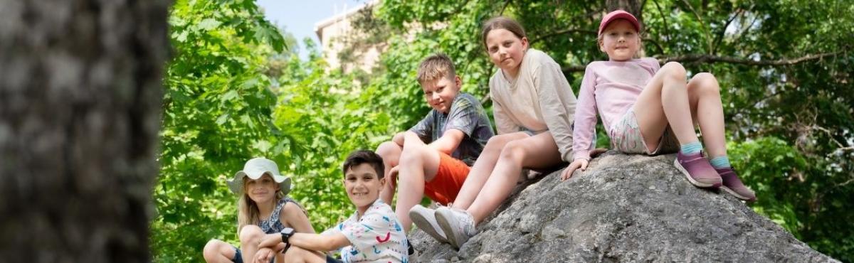 Viisi kouluikäistä lasta istuu kivellä vihreässä puistomaisemassa ja katsoo hymyillen kameraan. 