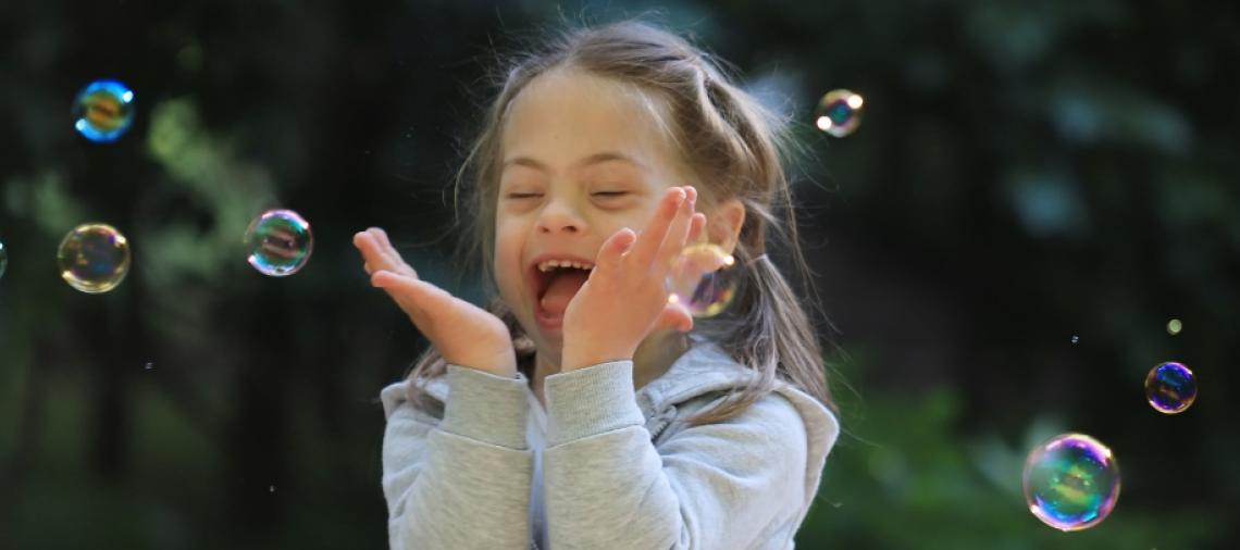 Pitkätukkainen, harmaaseen pusakkaan pukeutunut lapsi tavoittelee nauraen saippuakuplia. 